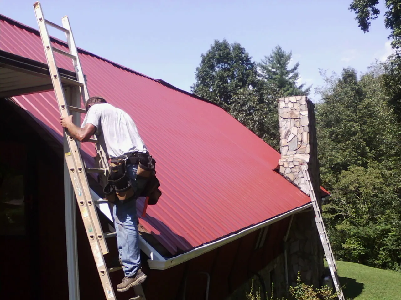Roofer on a ladder beside a red metal roof and stone chimney — Soffit & Fascia Repair by Roofing Brothers in Bretton Woods